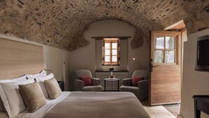 Et soveværelse med stenvæg, en seng med hvidt og beige sengetøj, to lænestole, et lille bord med en lampe og en trædør.