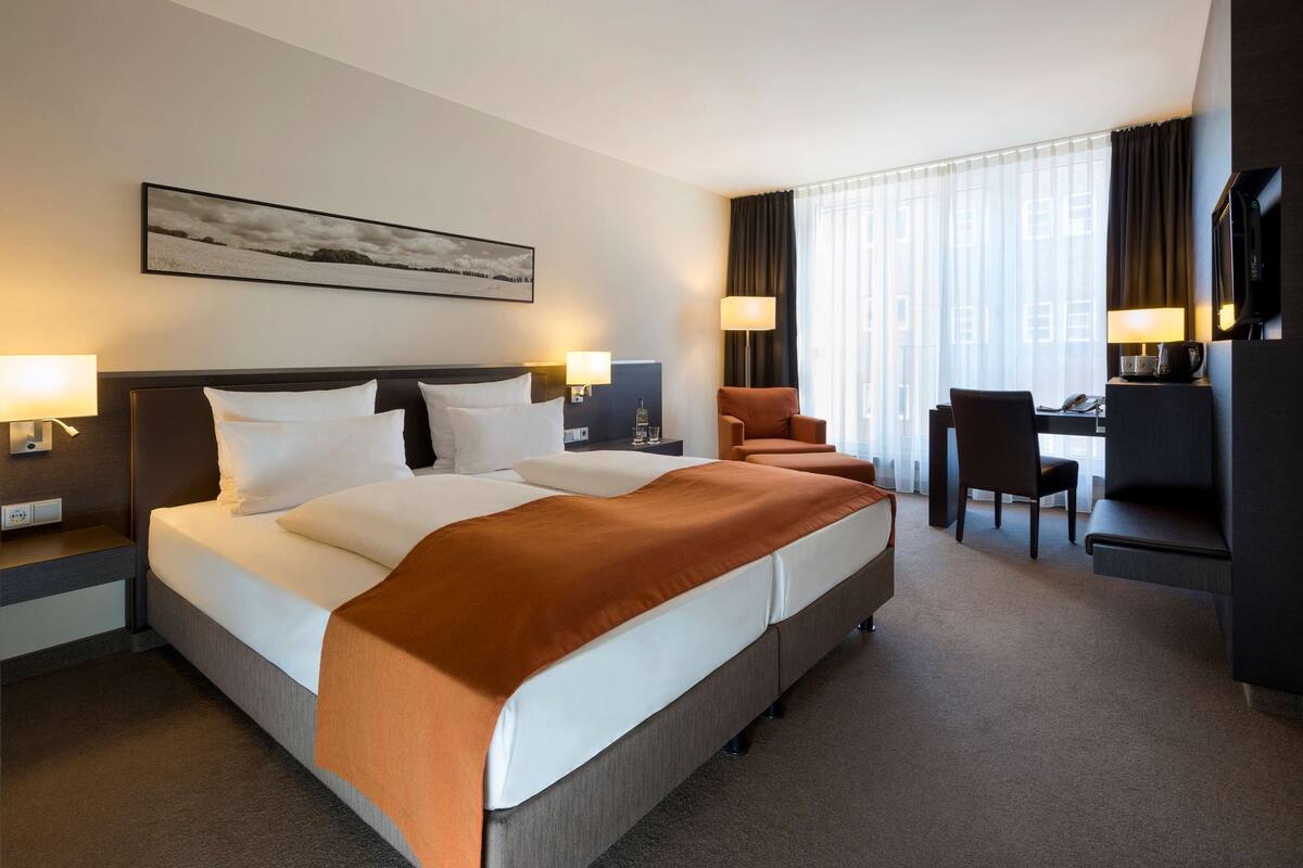 Une chambre d’hôtel avec un grand lit, deux tables de chevet, un bureau avec une chaise, une télévision et une fenêtre avec des rideaux.