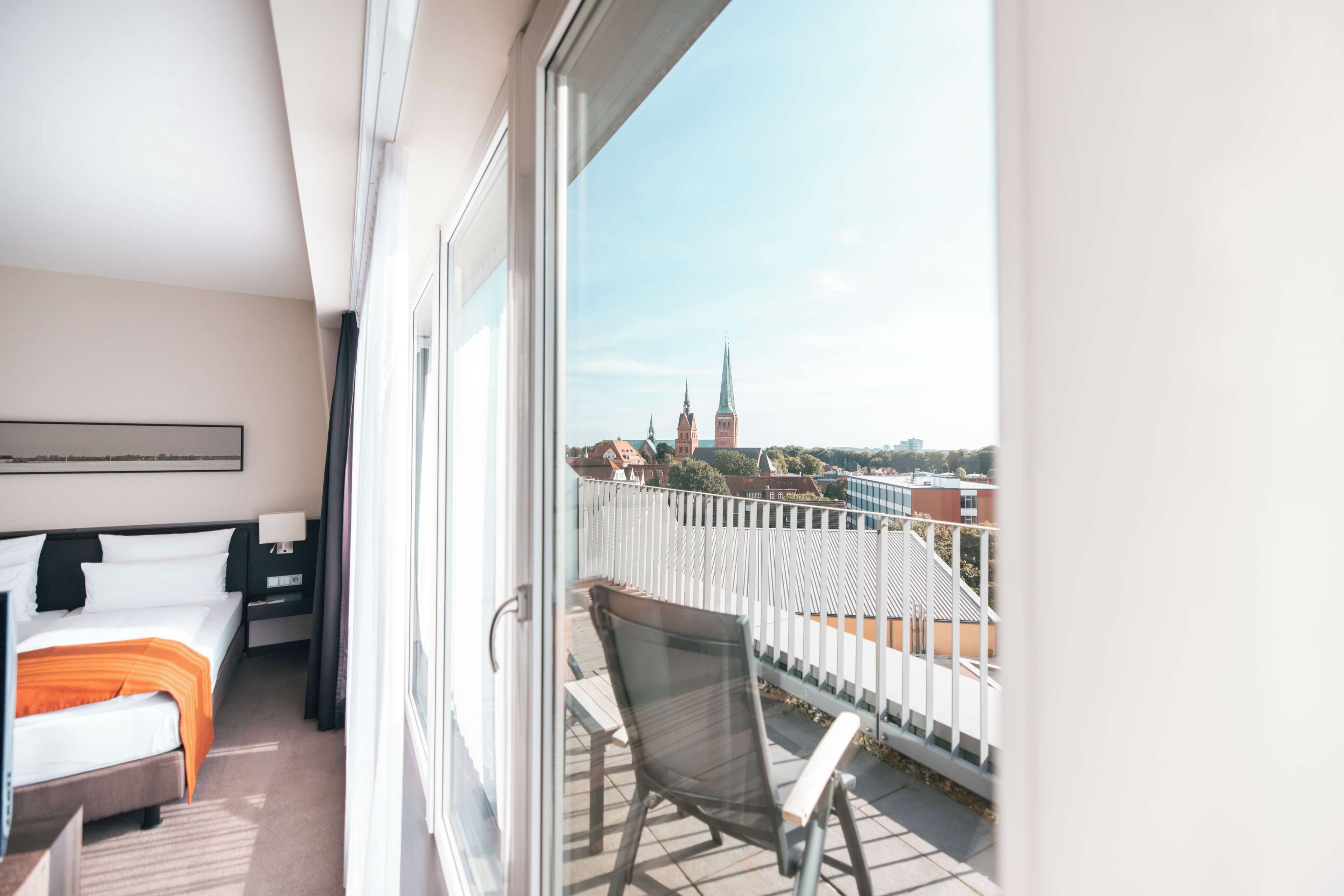 Ein Hotelzimmer mit Balkon, einem Bett, einem Sessel und Blick auf eine Stadtlandschaft mit einem Kirchturm.