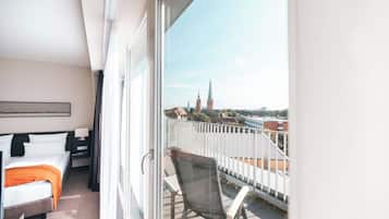 Ein Hotelzimmer mit Balkon, einem Bett, einem Sessel und Blick auf eine Stadtlandschaft mit einem Kirchturm.