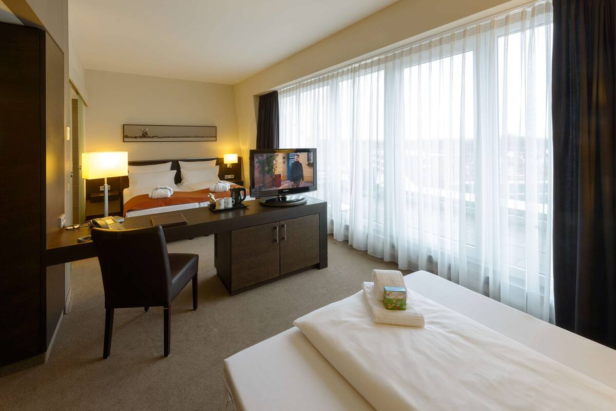 Une chambre d’hôtel avec un lit, un bureau équipé d’un ordinateur, une télévision et une fenêtre avec des rideaux légers.