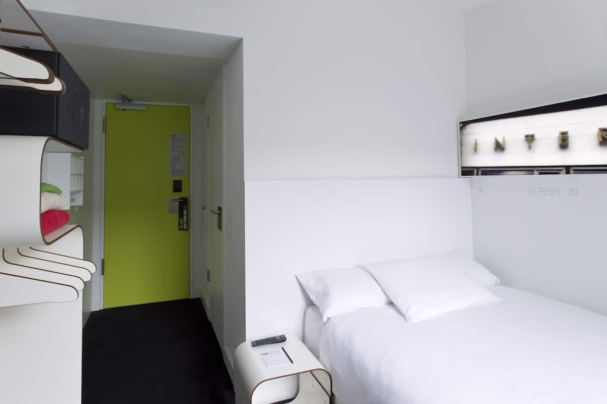 A hotel room with a bed, a bedside table, a green door, and a white wall with a light fixture.