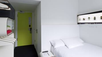 Habitación de hotel con cama, mesita de noche, puerta verde y pared blanca con lámpara.