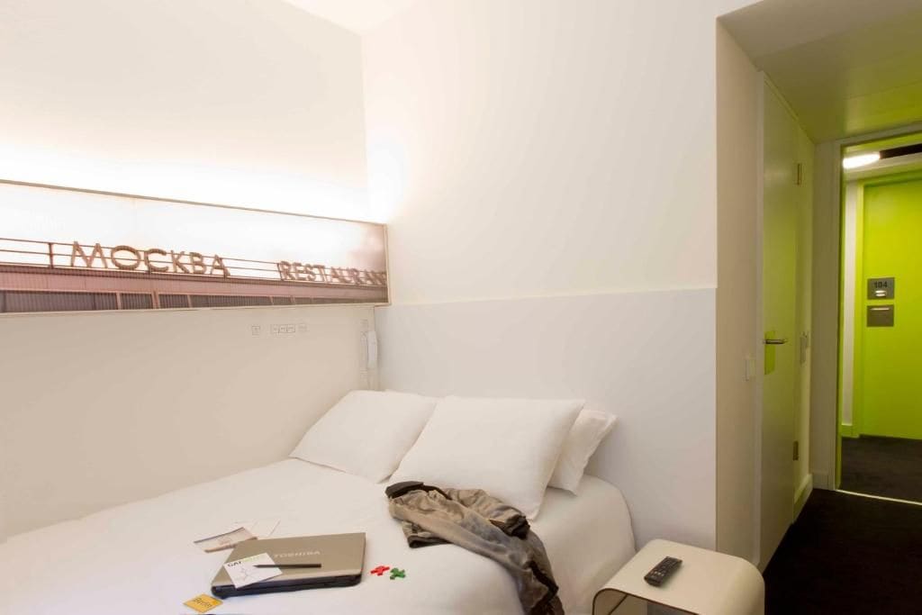 A hotel room with a bed, a bedside table with a remote control, and a wall-mounted sign above the bed.