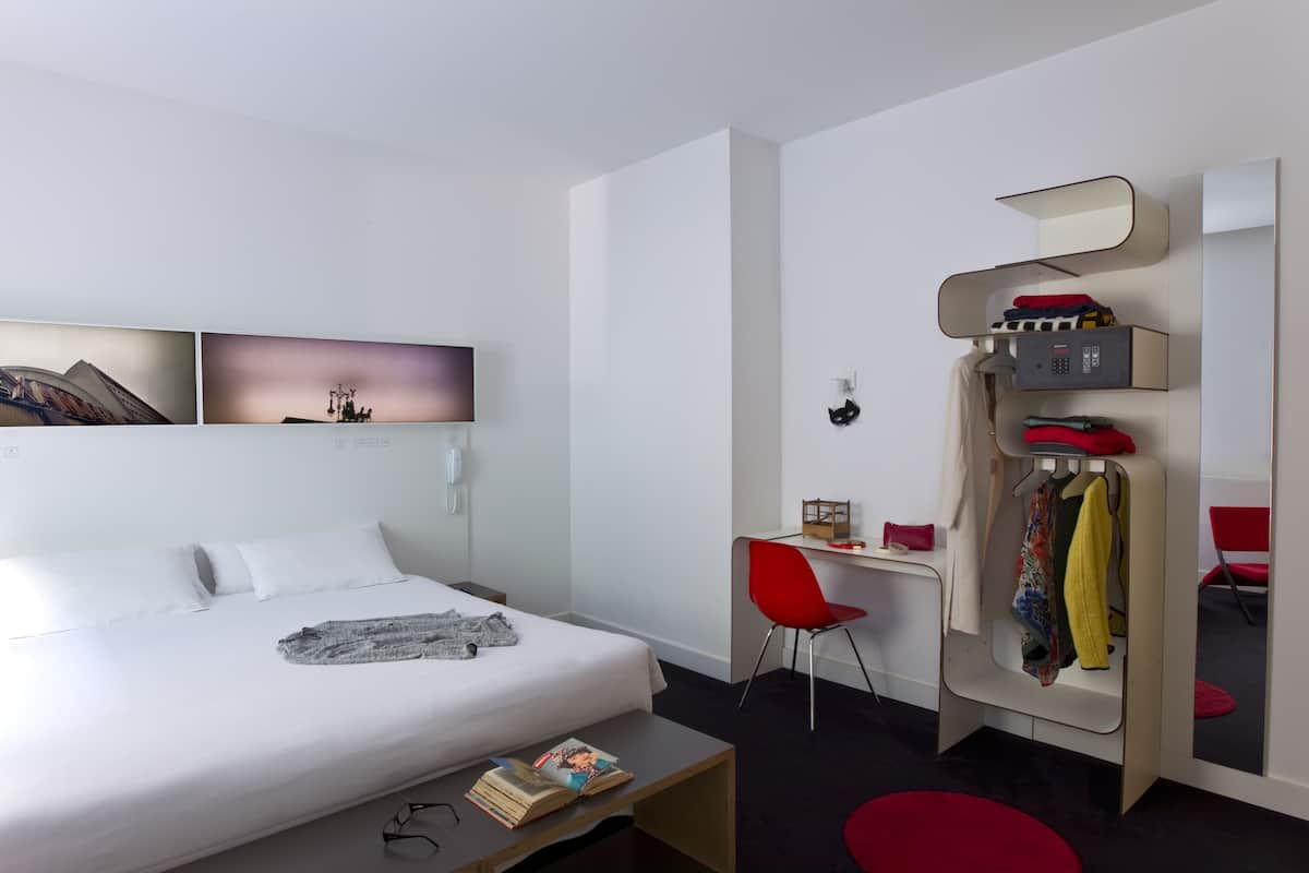 A modern hotel room with a bed, a desk, a chair, and a wardrobe.
