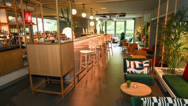 A modern bar area with wooden furniture, a counter with various items, and a seating area with green sofas and a round table. - Hotel Gat Point Charlie Berlin (Berlin)