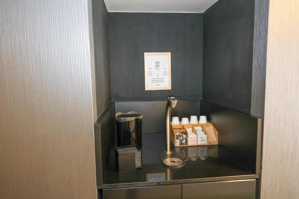 A small, enclosed area with a coffee machine, a trash bin, and a shelf with coffee cups and a box of tissues.