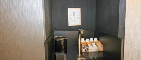 A small, enclosed area with a coffee machine, a trash bin, and a shelf with coffee cups and a box of tissues.