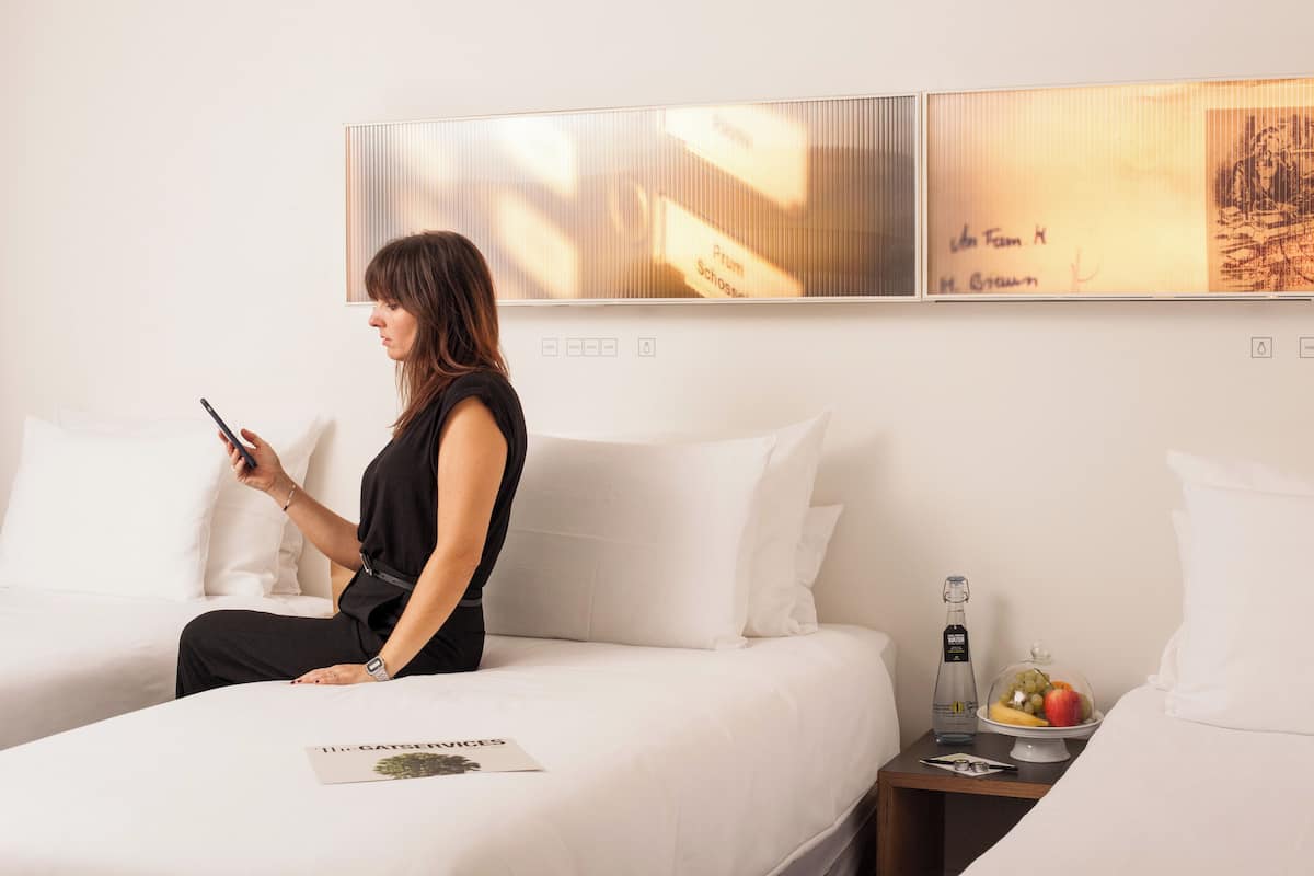 A hotel room with a bed, a nightstand with a bottle and fruit, and a wall with artwork.
