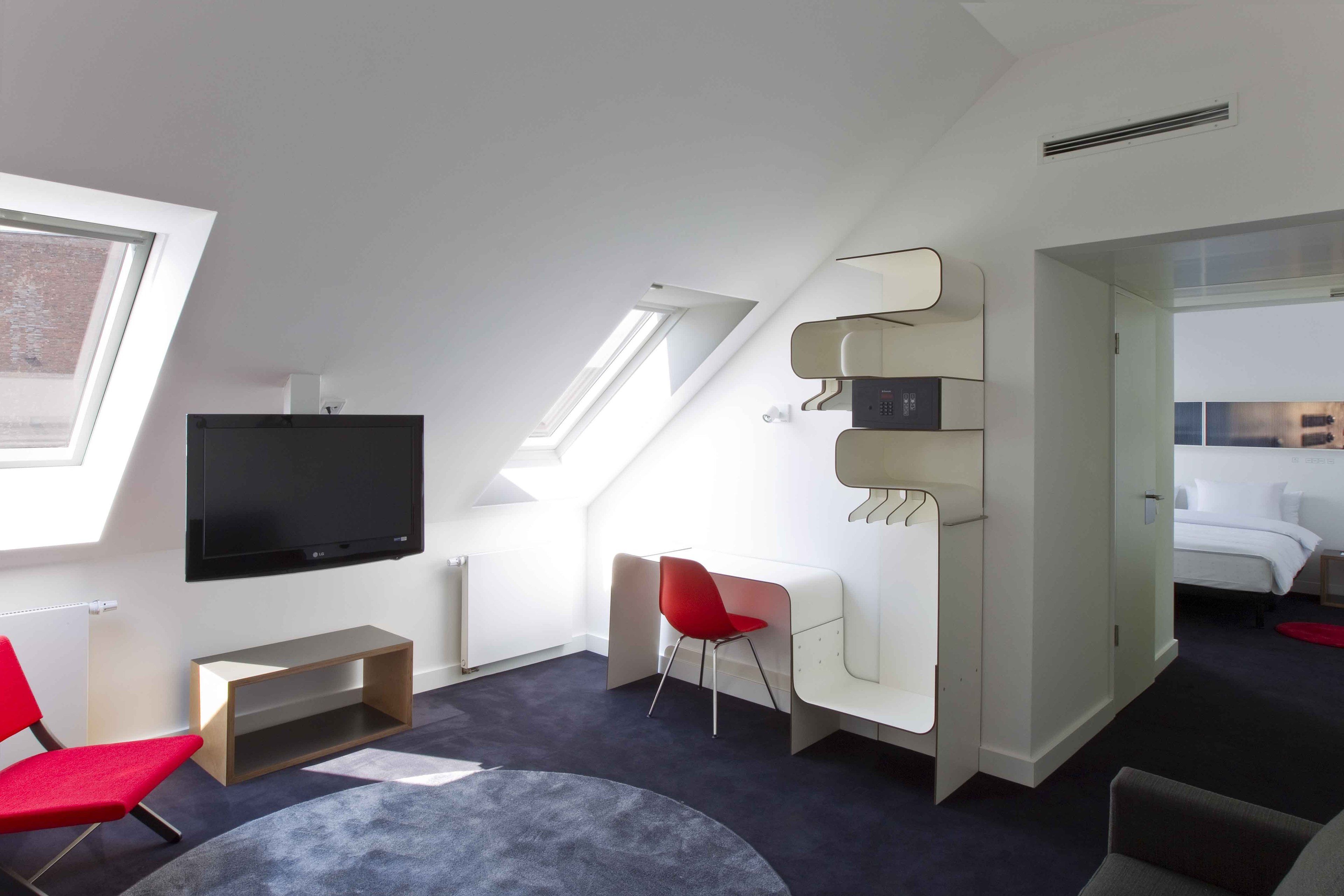 A modern, well-lit room with a flat-screen TV, red chairs, a small table, and a bed with white bedding.