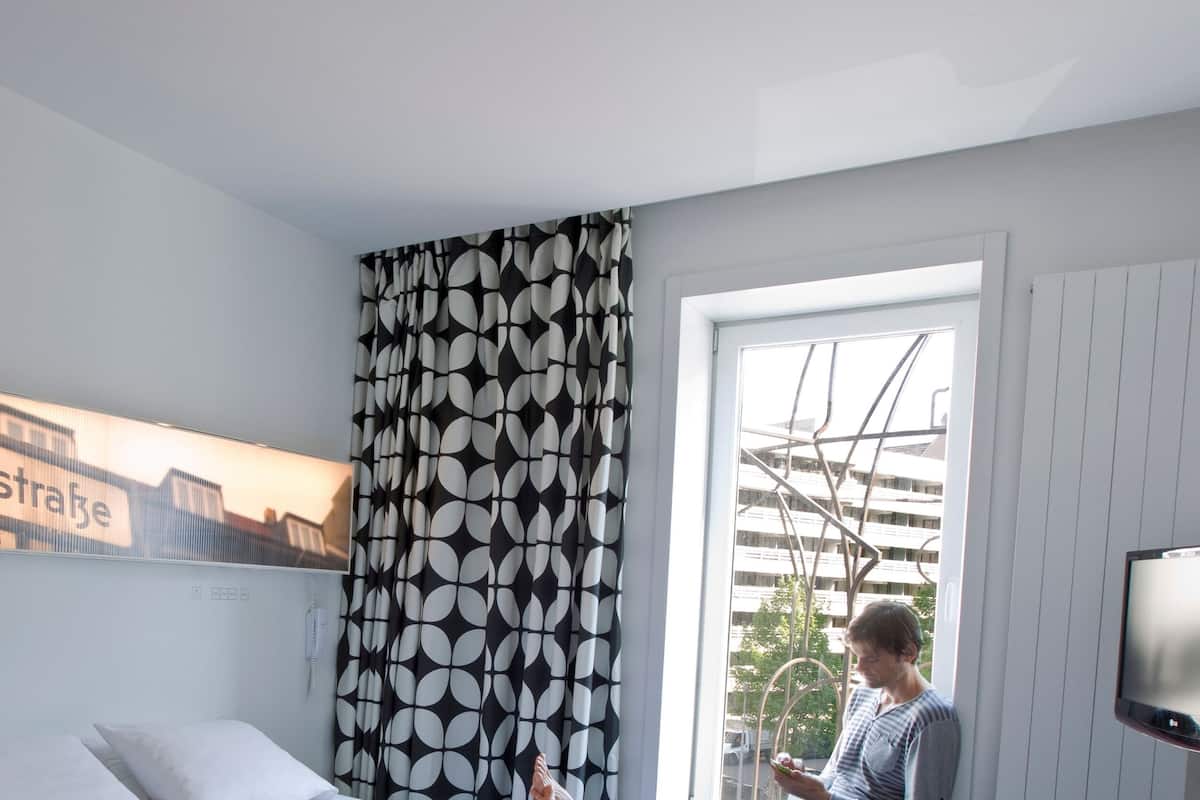 A hotel room with a bed, a window with a view of buildings, a curtain with a leaf pattern, and a sign on the wall.