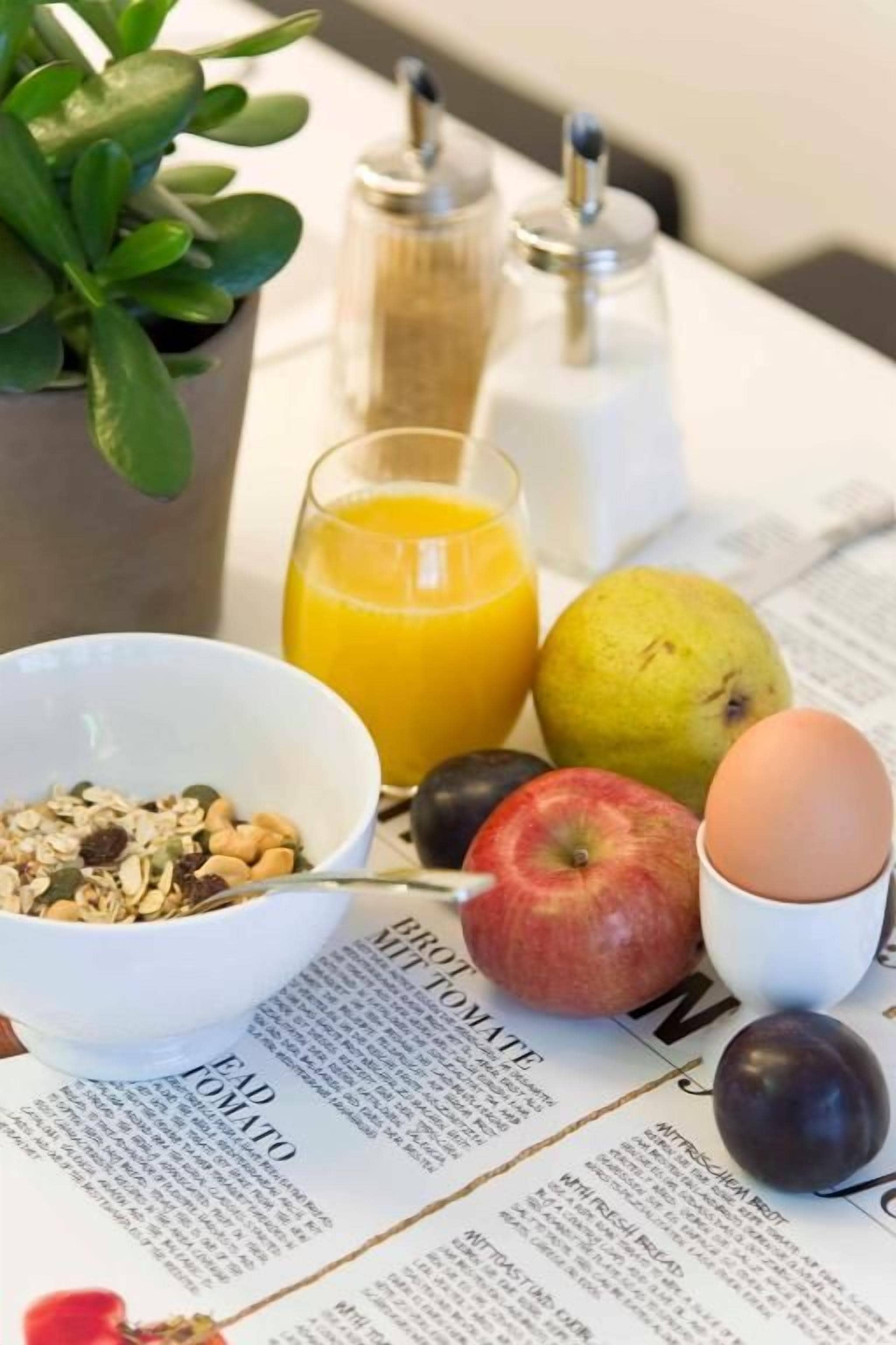 A breakfast setting with a bowl of cereal, a glass of orange juice, an apple, a plum, and a boiled egg on a table with a newspaper.