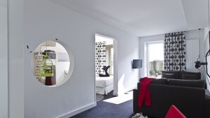 A modern hotel room with a round mirror, a bed with a geometric-patterned curtain, a grey sofa, a red chair, and a TV mounted on the wall. - Hotel Gat Point Charlie Berlin (Berlin)