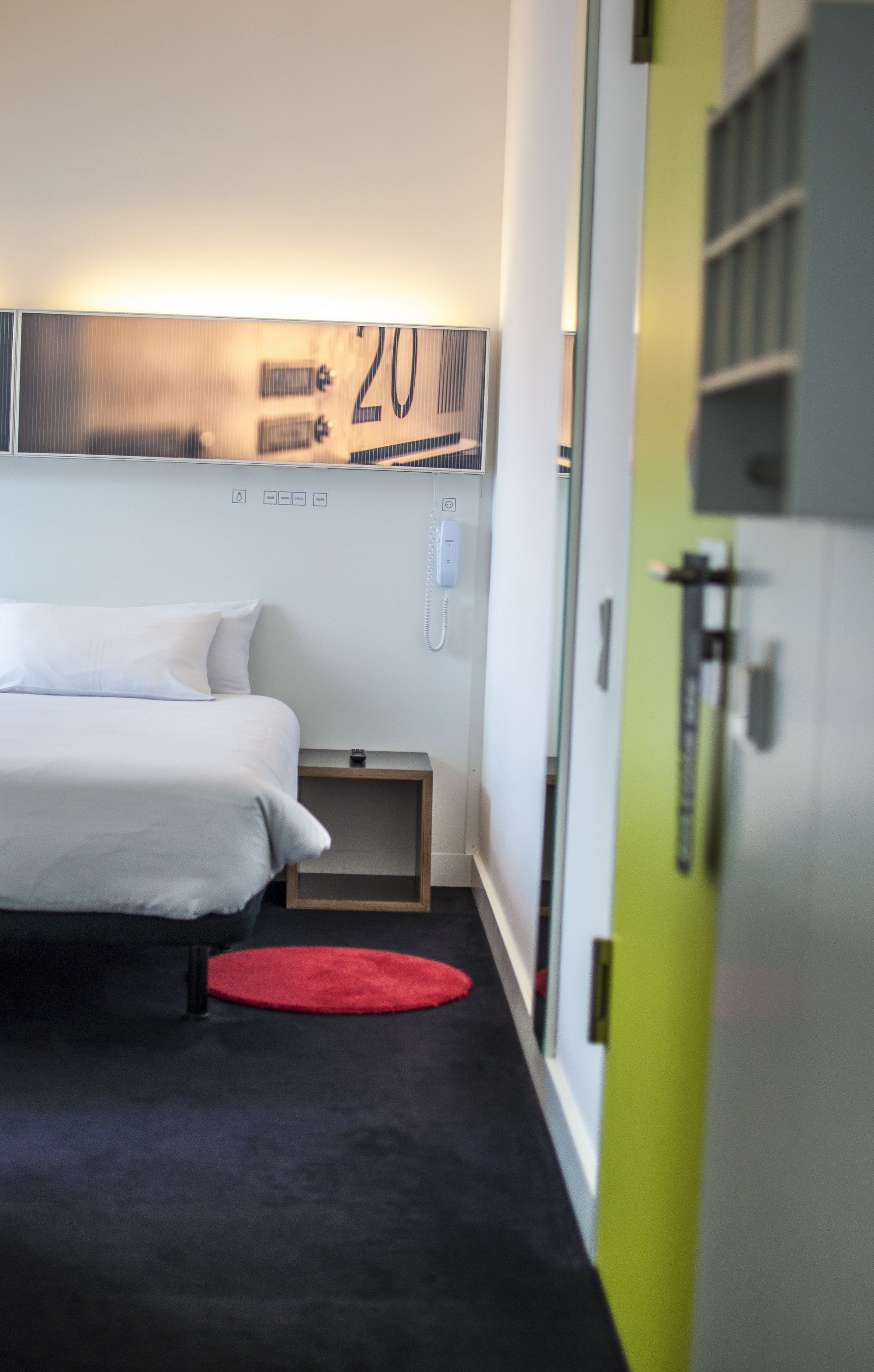 Hotelkamer met een bed, een rood tapijt, een nachtkastje, een telefoon en een deur met een nummerbord.