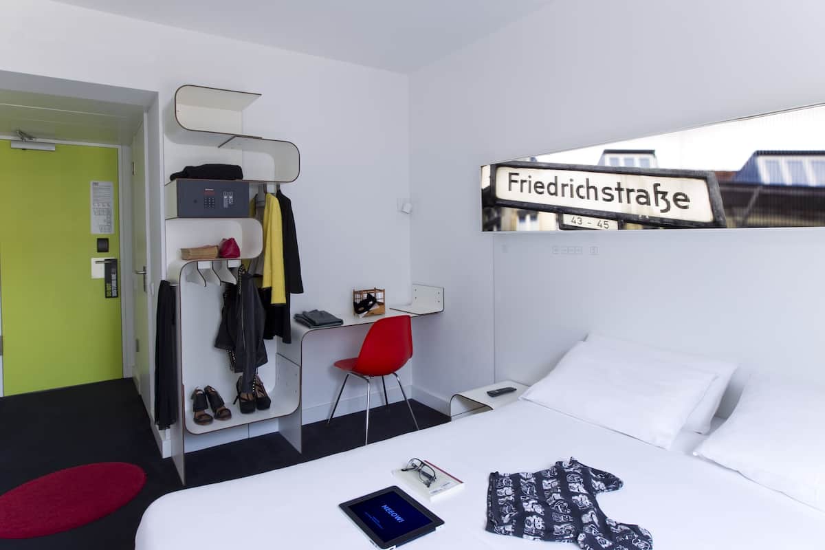 A hotel room with a bed, a desk with a chair, a coat rack, and a sign indicating the street name.