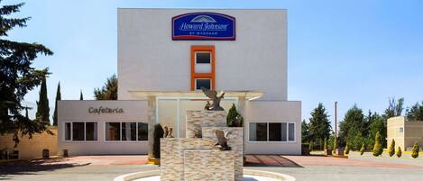 A Howard Johnson by Wyndham hotel with a fountain in front, a sign for the cafeteria, and a clear blue sky.