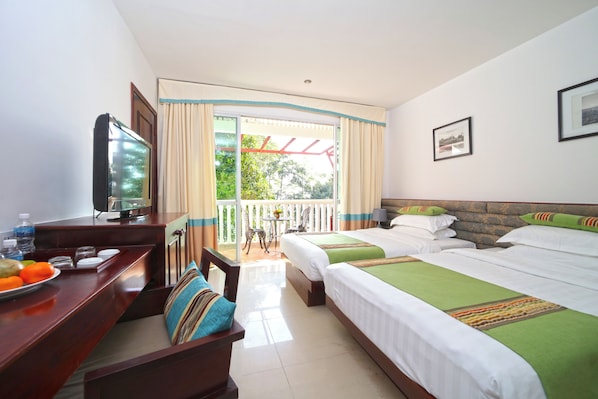 Habitación de hotel con dos camas, un escritorio con televisor, un balcón con mesa y sillas, y vista a zonas verdes.