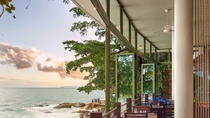 A restaurant with wooden tables and chairs, set against a backdrop of the ocean and rocky shoreline. - Independence Hotel by Dara (Sihanoukville)