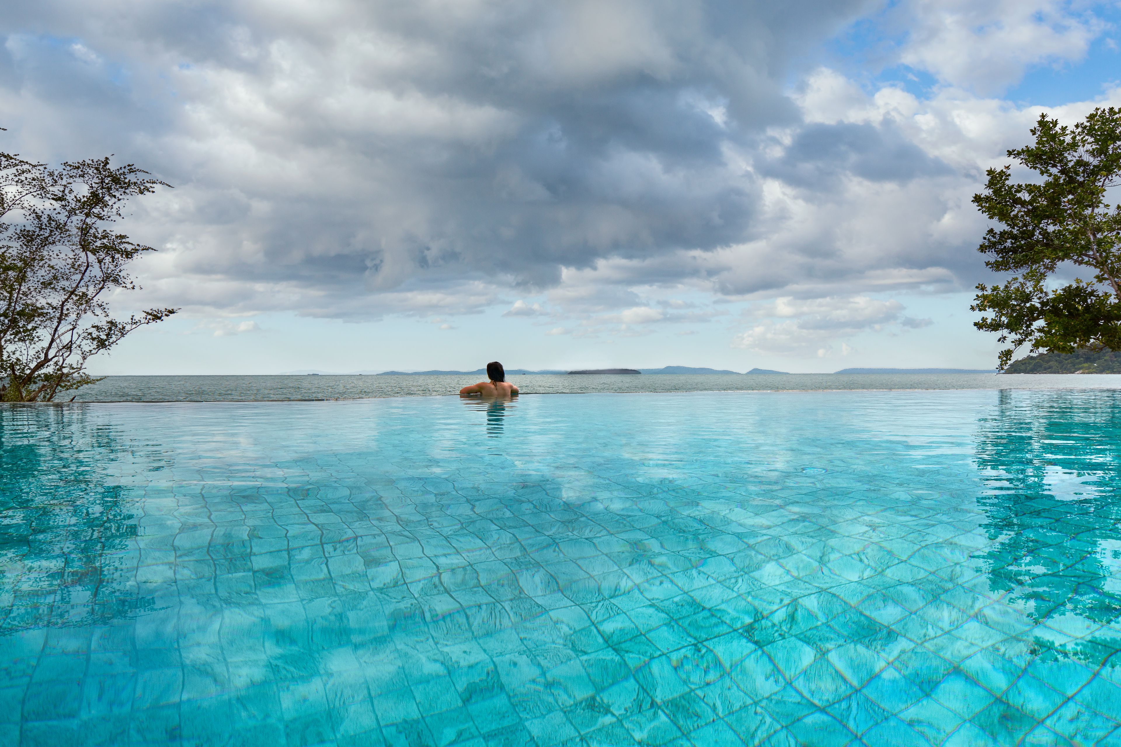 A person swimming in a clear blue pool with a view of distant mountains and a cloudy sky.