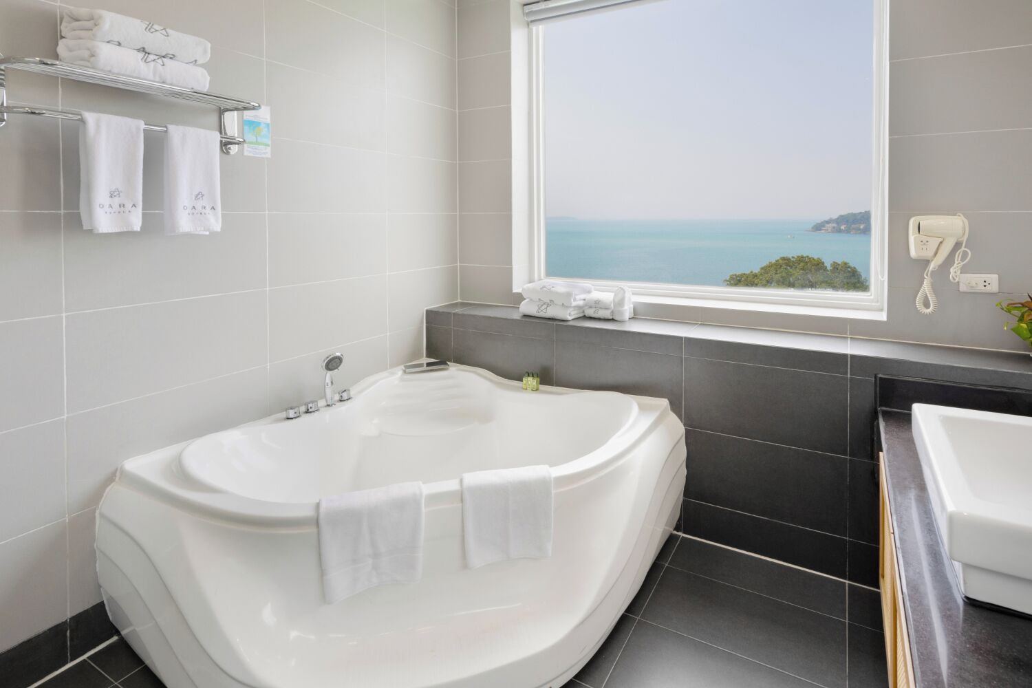 A modern bathroom with a large freestanding bathtub, a window with a view of the sea, and a vanity with a mirror and sink.