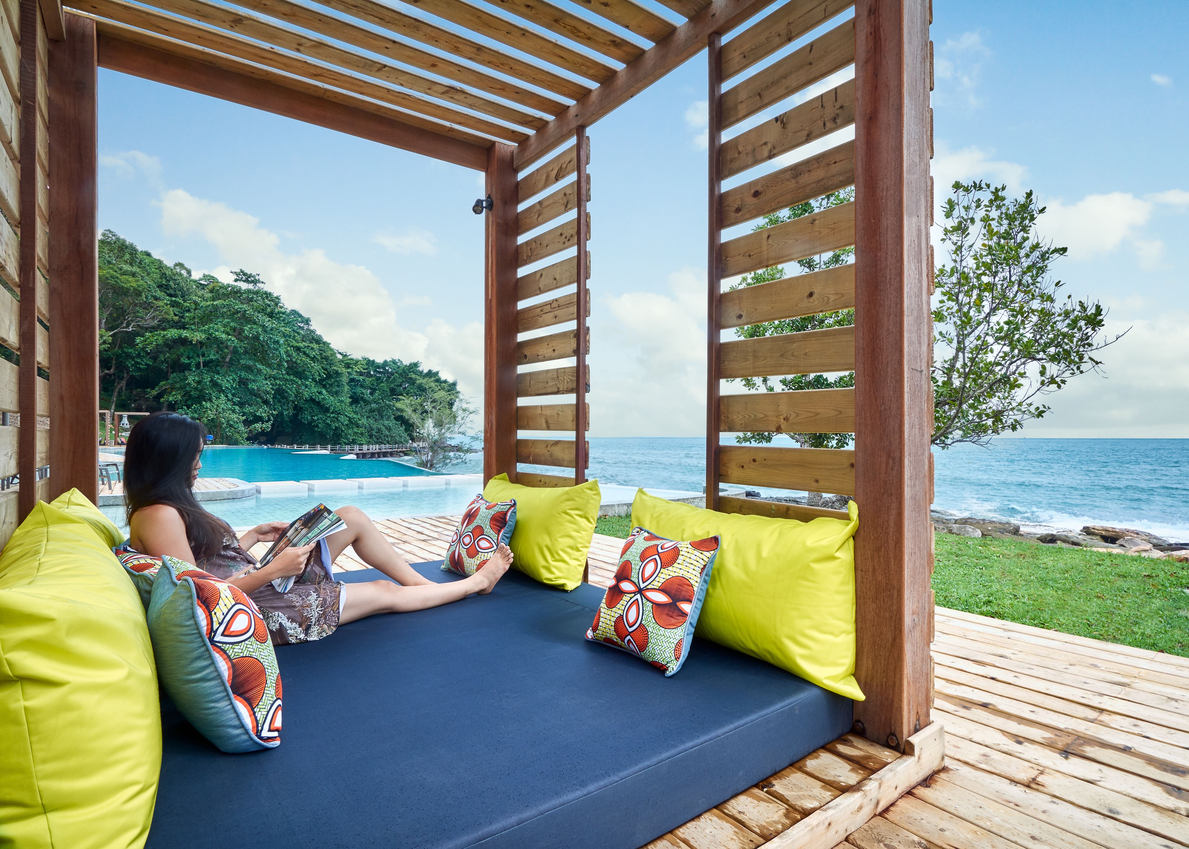 Une pergola en bois avec un coin salon agrémenté de coussins bleus, une piscine et une vue sur l’océan.