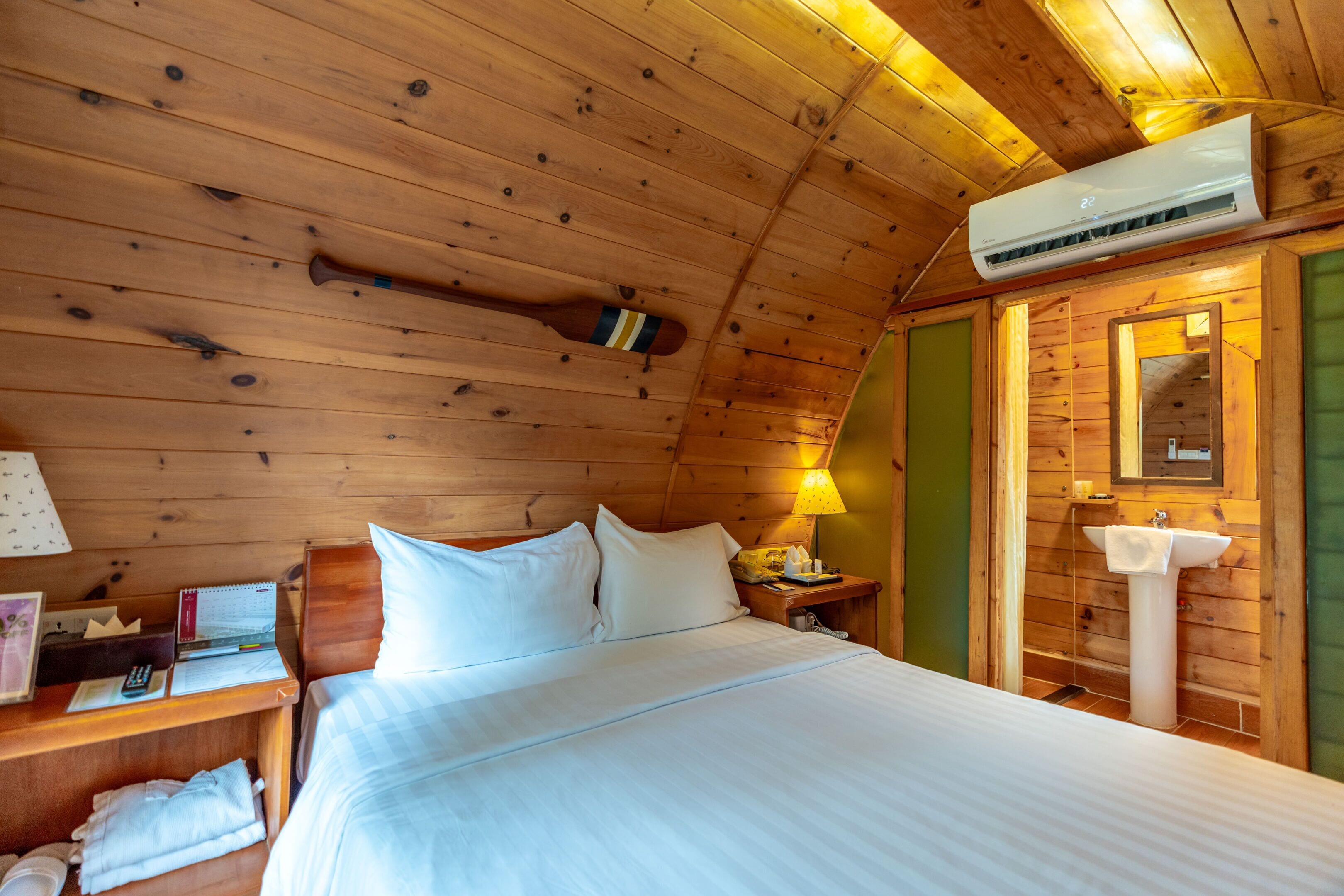 A wooden cabin room with a bed, a nightstand, a lamp, a mirror, and an air conditioning unit.