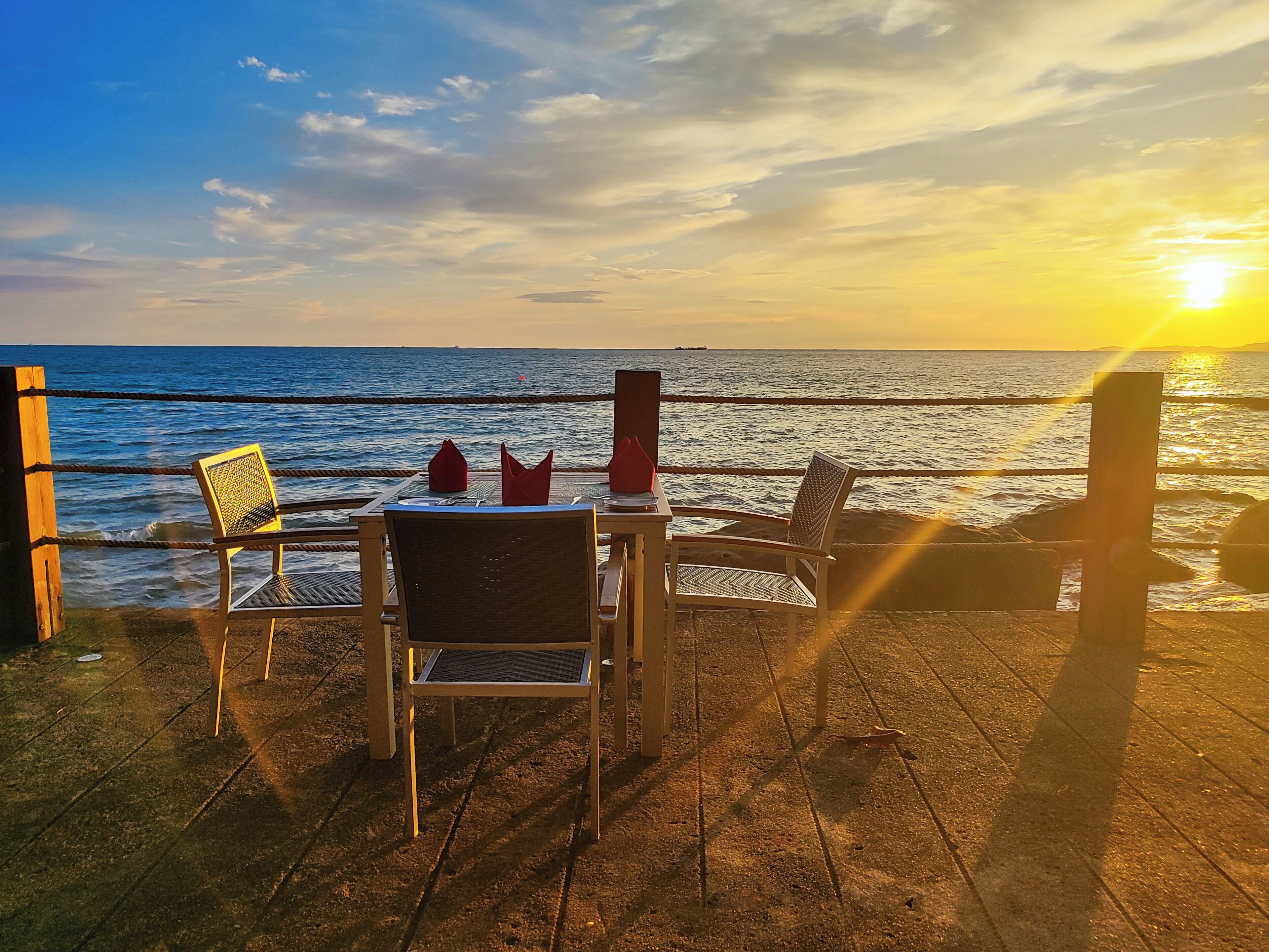 A table and chairs on a wooden deck overlooking the sea with a sunset in the background.