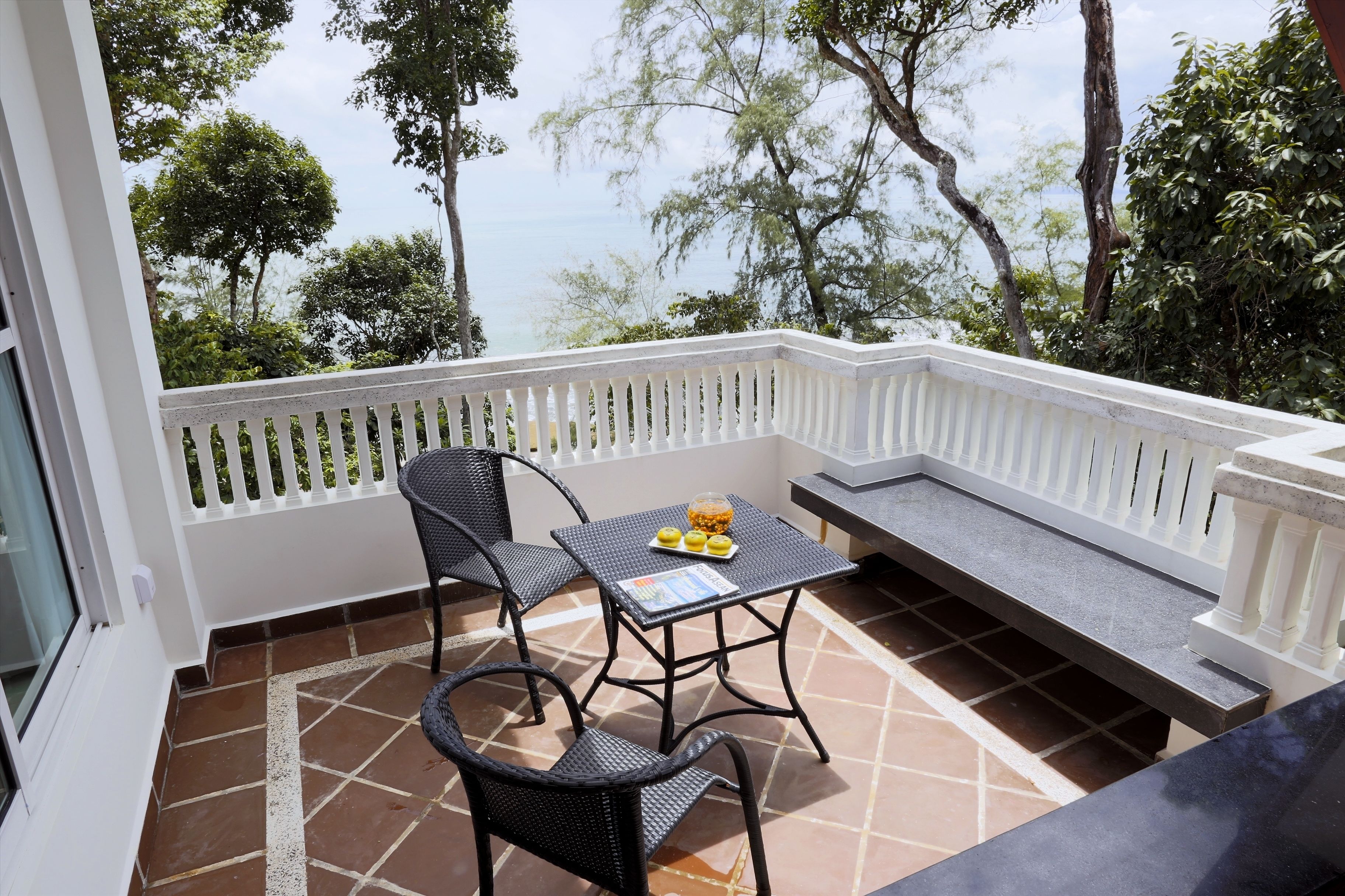 A balcony with a table and chairs, a bench, and a view of trees and the sea.