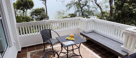 A balcony with a table and chairs, a bench, and a view of trees and the sea.