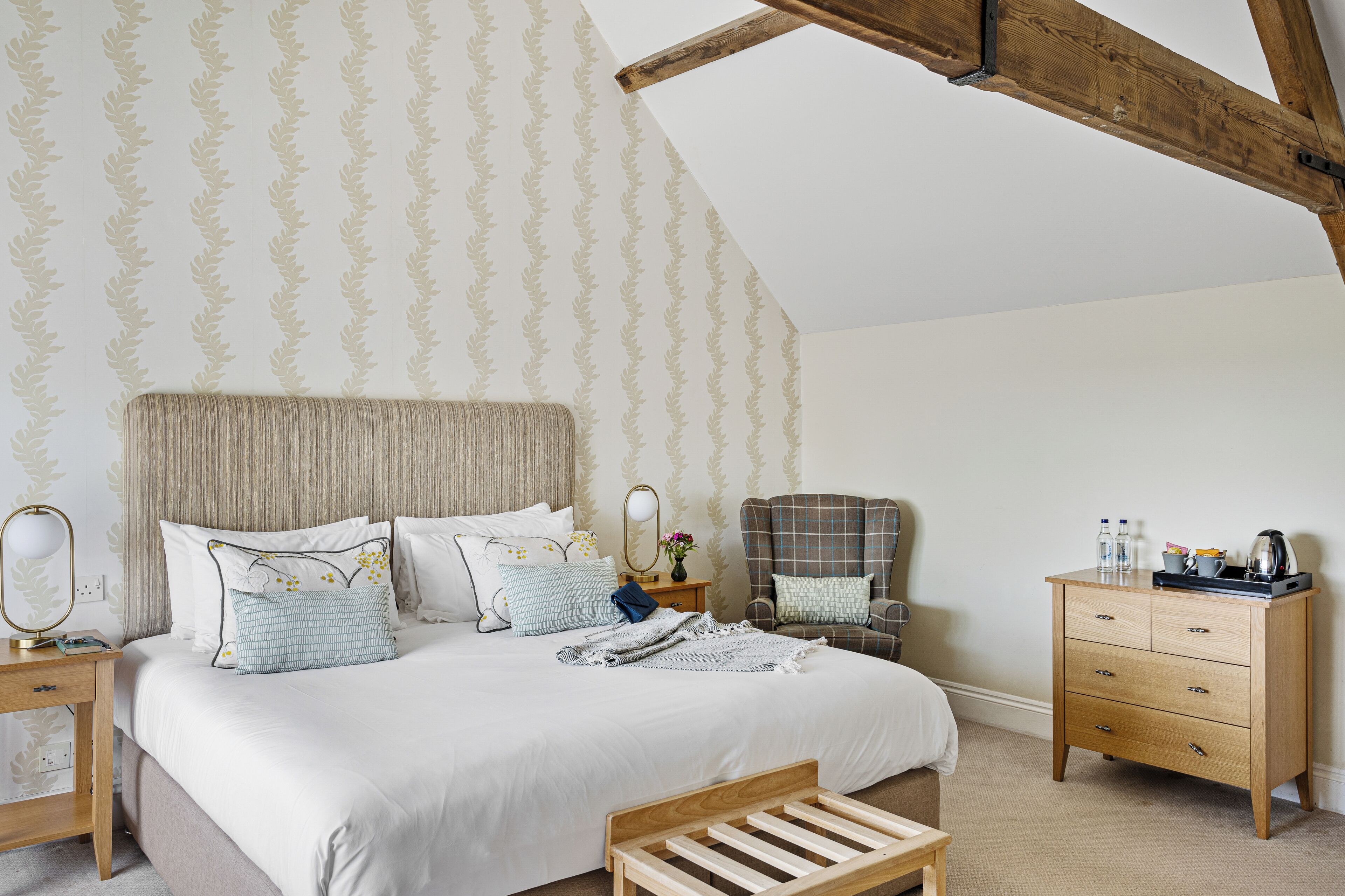 A bedroom with a large bed, a nightstand, a chair, a wooden dresser, and a wooden bench.