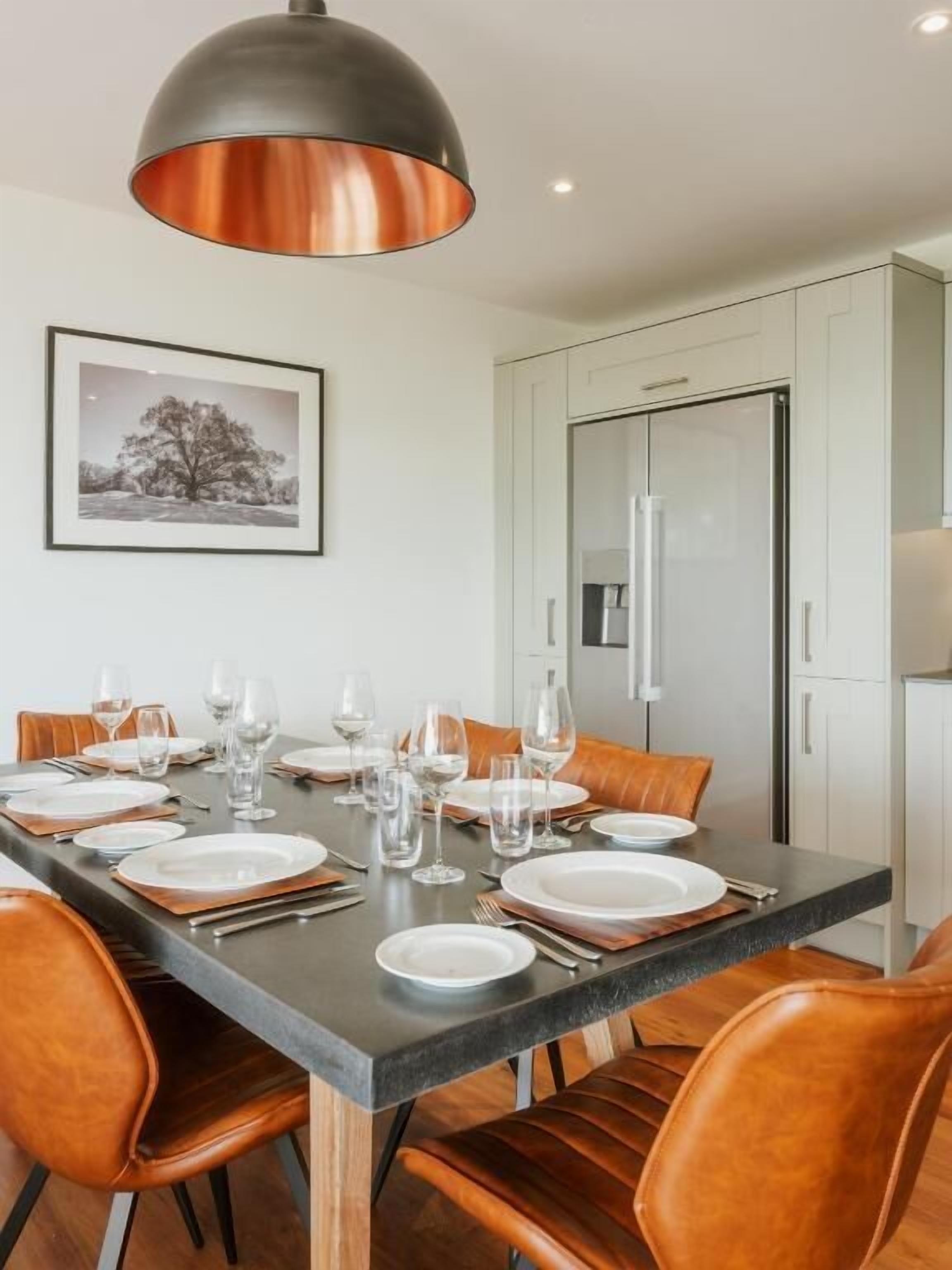 A dining area with a table set for six, surrounded by leather chairs, a modern pendant light, and a framed picture on the wall.