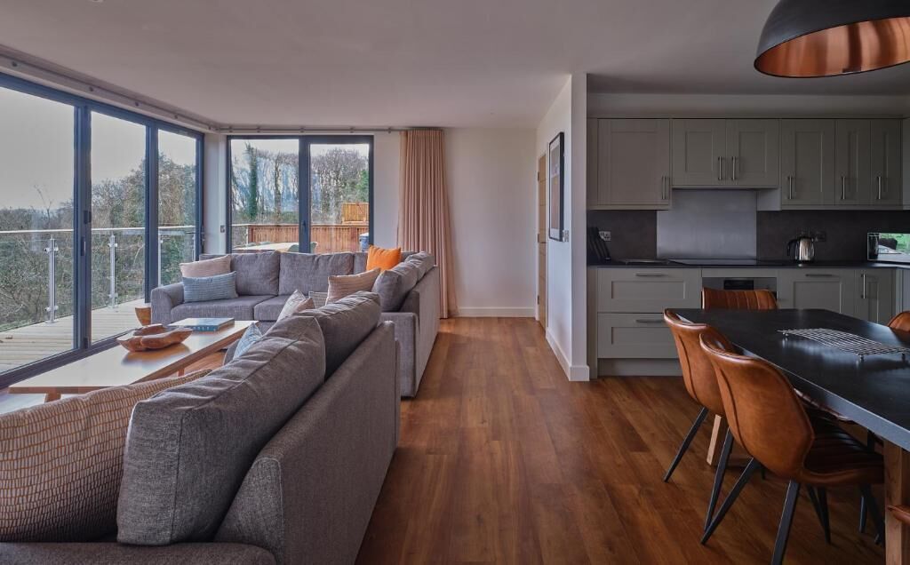 A modern living area with a sofa set, dining table, and chairs, a kitchen area with cabinets, and large windows offering a view of the outdoors.