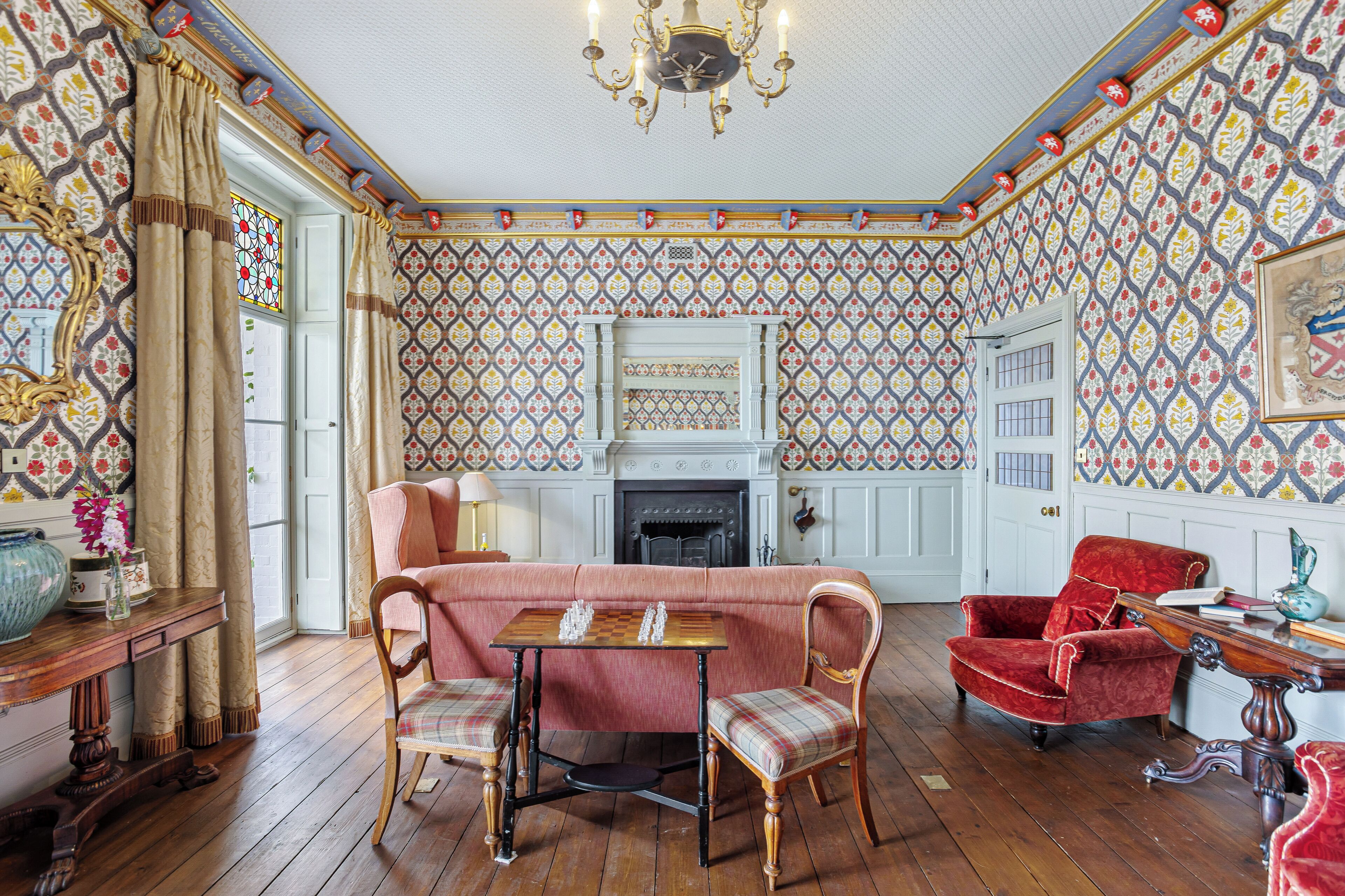 Una habitación con papel tapiz estampado, una chimenea, un sillón rojo, una mesa de madera con piezas de ajedrez y una araña.