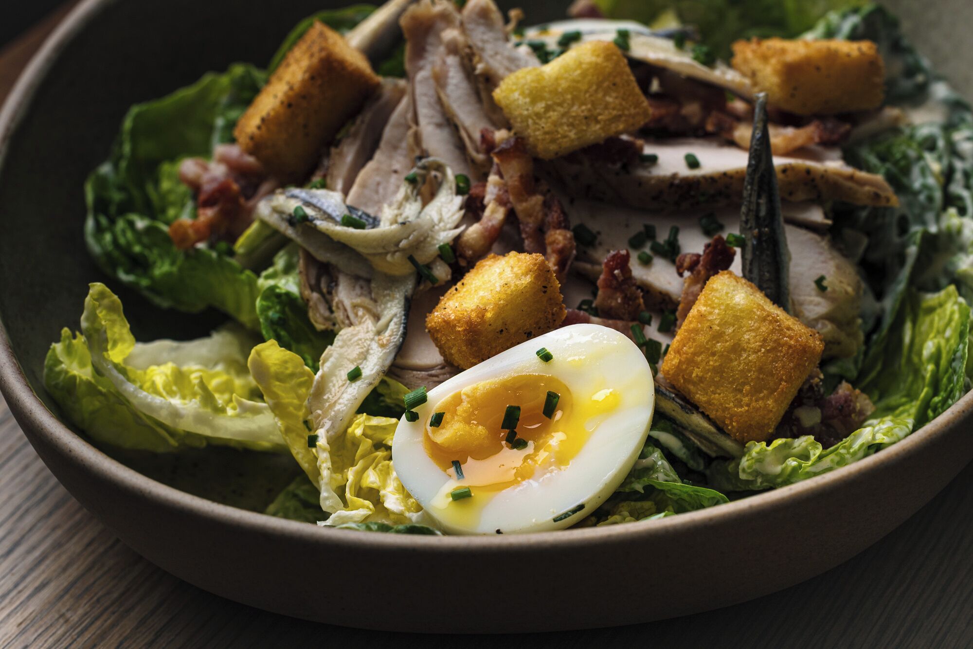A bowl of salad with lettuce, croutons, sliced meat, and a half-boiled egg.