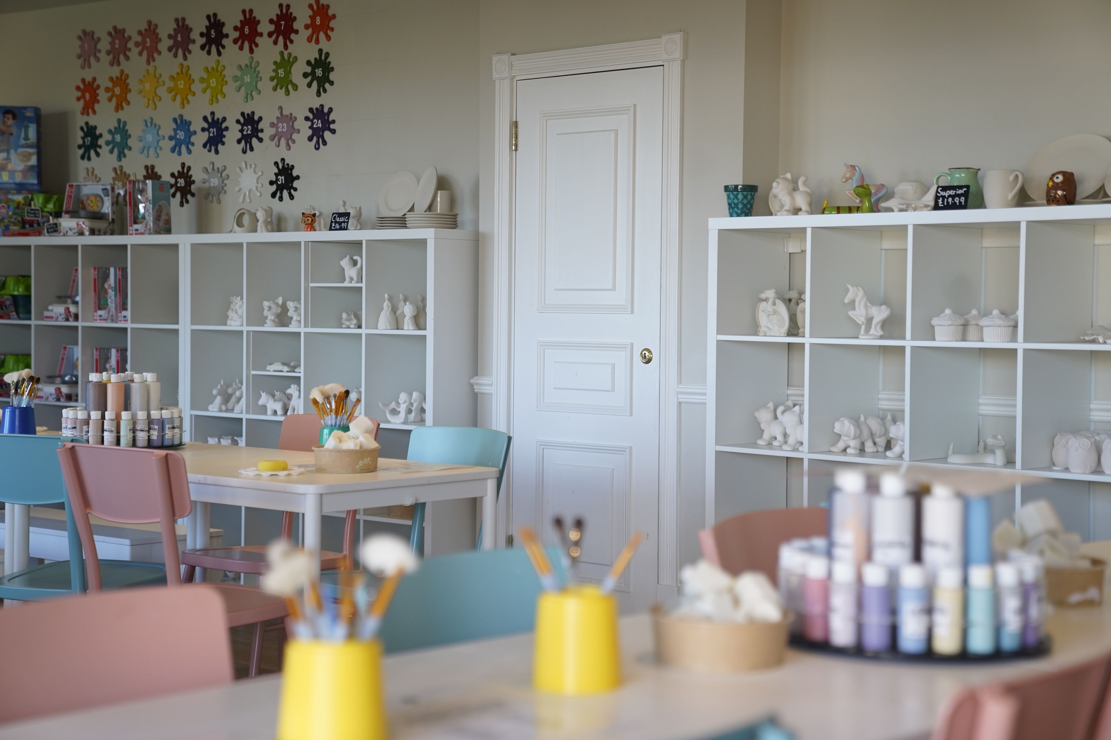 Une pièce avec des étagères blanches remplies d’objets décoratifs, une porte blanche et une table avec du matériel de dessin et de peinture.