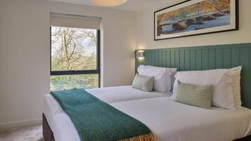 A bedroom with two beds, a green headboard, a painting of a landscape, and a window with a view of trees.