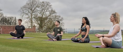 Four people sitting on yoga mats in an outdoor setting with a grassy field and trees in the background.