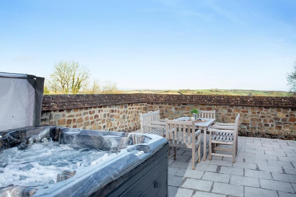 A hot tub with a stone wall backdrop, wooden chairs, and a table on a paved patio.
