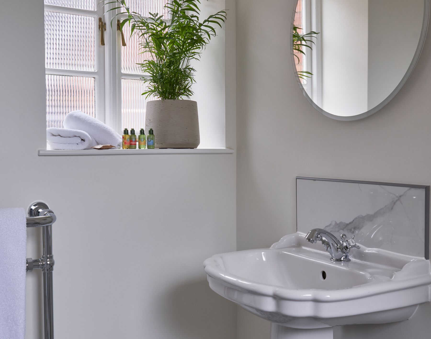 Een badkamer met een witte wastafel, een spiegel, een raam met een plant en een handdoekhouder.