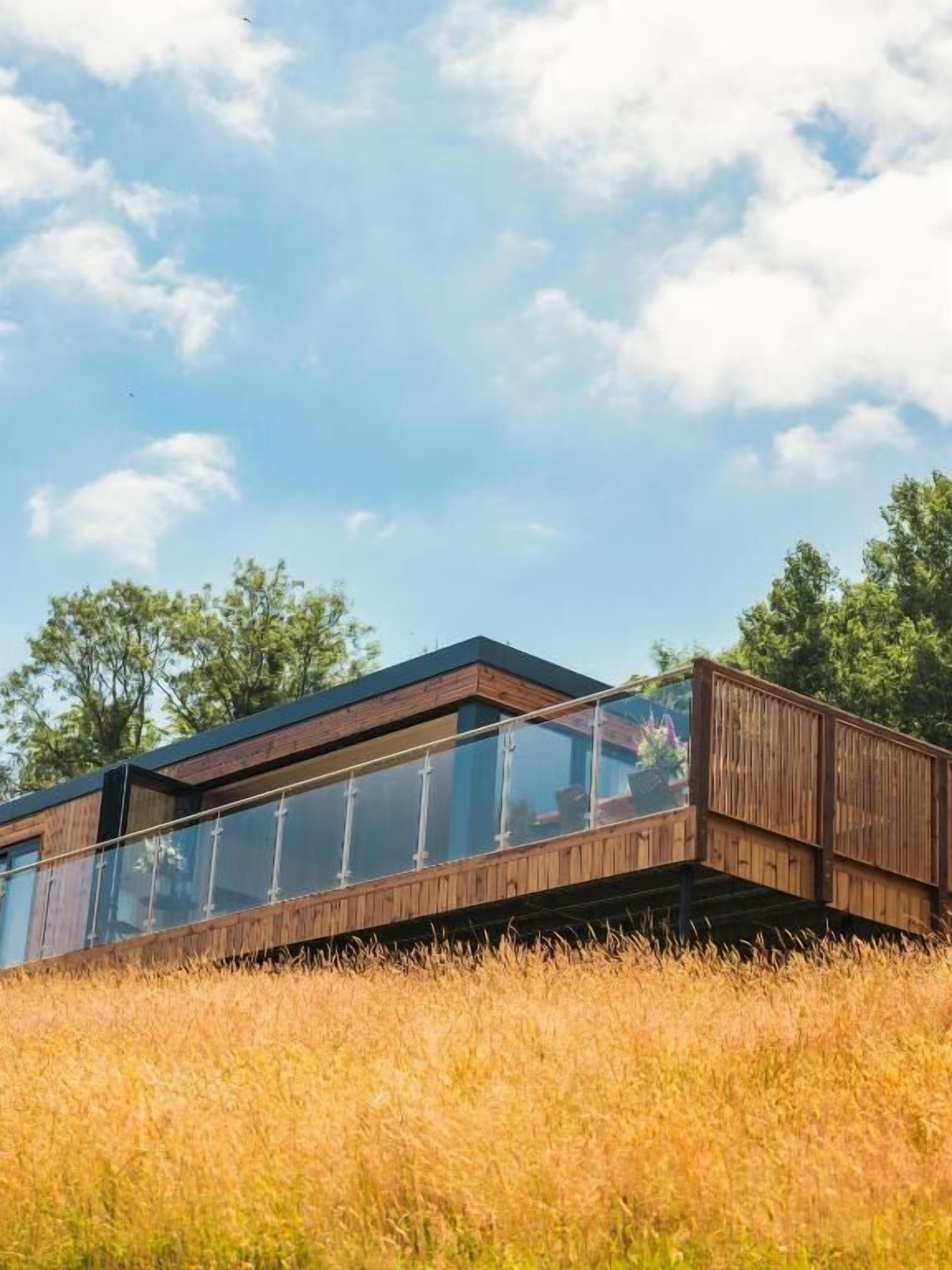 Een modern houten gebouw met glazen balustrades, gelegen in een grasveld met bomen op de achtergrond.