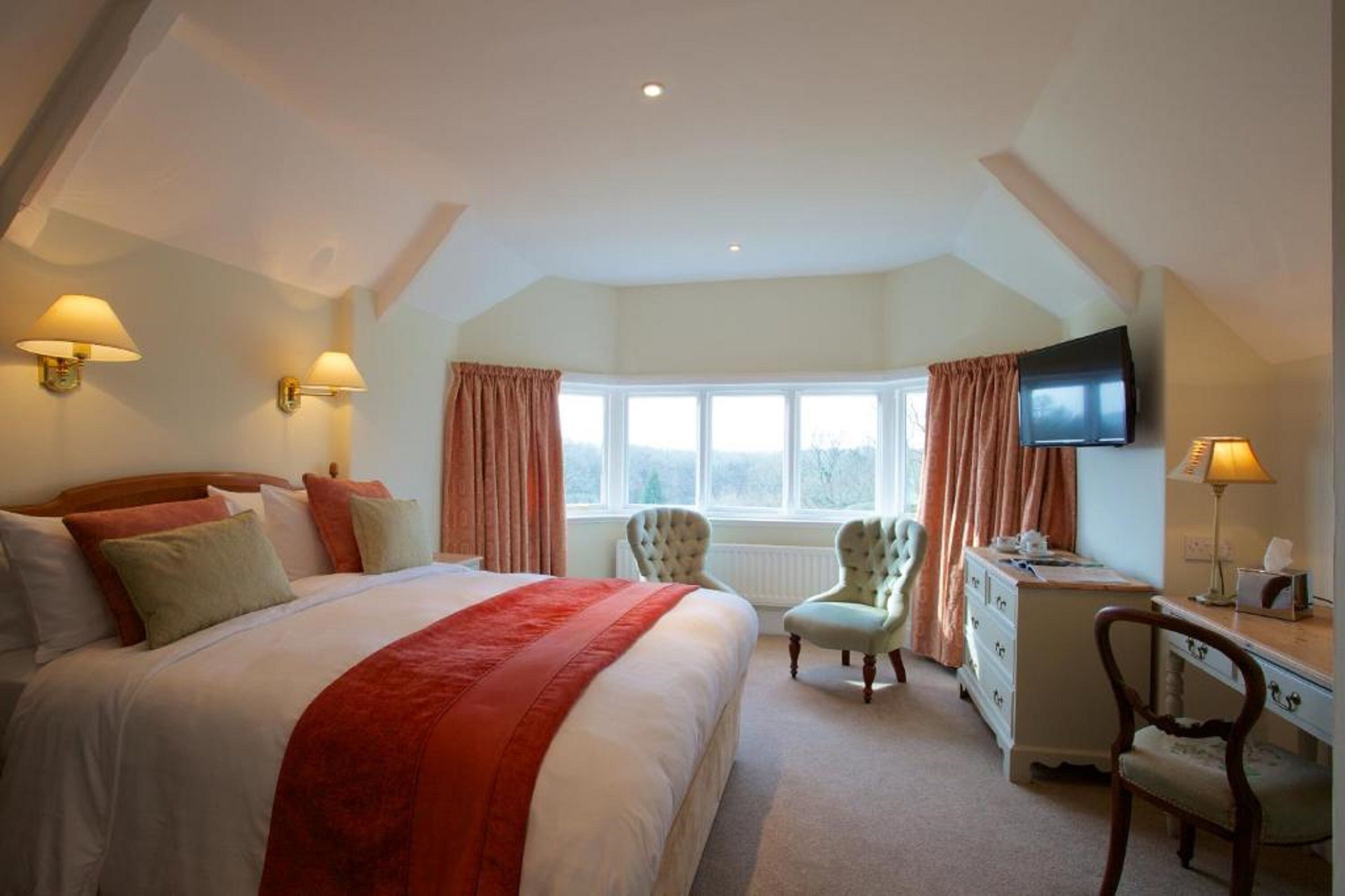 A bedroom with a large bed, a dresser, a desk, two chairs, a television, and a window with curtains.