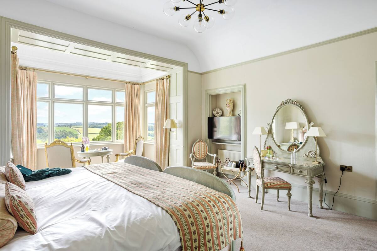Une chambre avec un grand lit, une coiffeuse avec un miroir, une chaise et une vue sur l’extérieur.