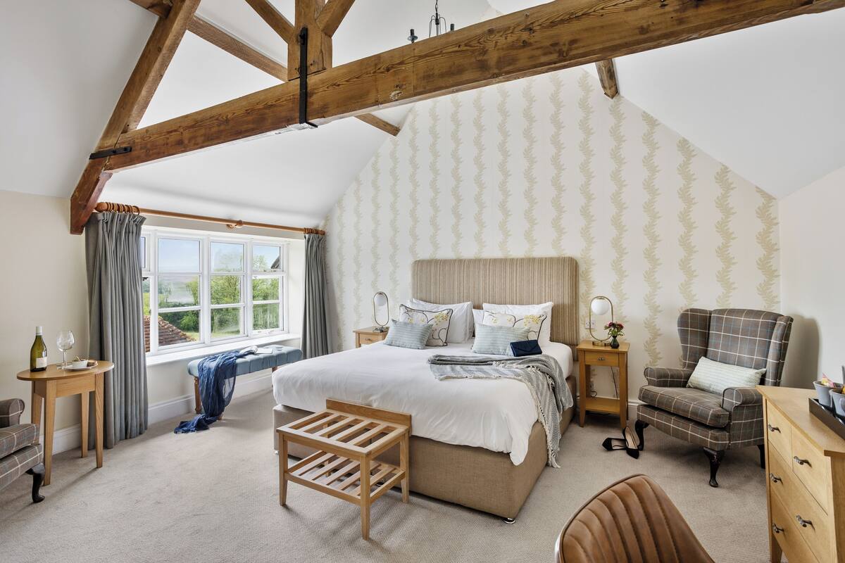 Une chambre à coucher avec un grand lit, un banc en bois, une chaise, un bureau et une fenêtre avec des rideaux.