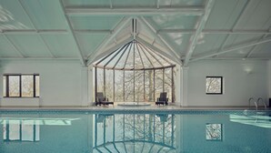 Una amplia piscina cubierta con techo de cristal, dos sillones de descanso y vistas a árboles a través del techo.