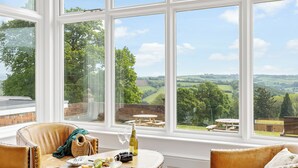 Habitación con un amplio ventanal que ofrece vistas panorámicas al campo. Hay una mesa redonda de madera con una botella de vino y un plato de comida. Dos sillas tapizadas están dispuestas alrededor de la mesa.