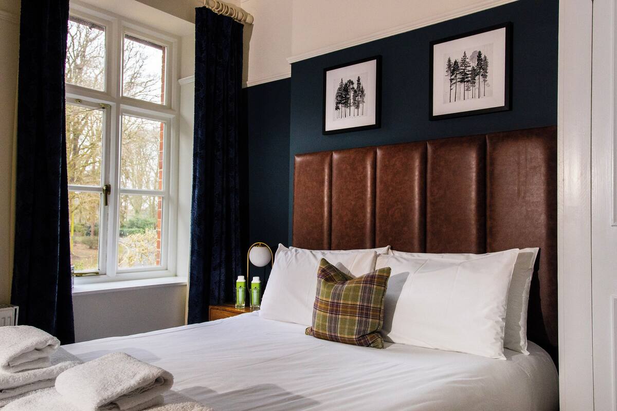 Un lit bien fait, avec du linge de lit blanc et un coussin à carreaux, une table de chevet avec une lampe, et deux tableaux encadrés accrochés au mur.