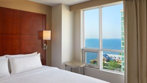A hotel room with a large bed, a small round table, a nightstand, a lamp, and a window with a view of the sea and buildings. - Residence Inn by Marriott Kingston Water's Edge (Kingston)