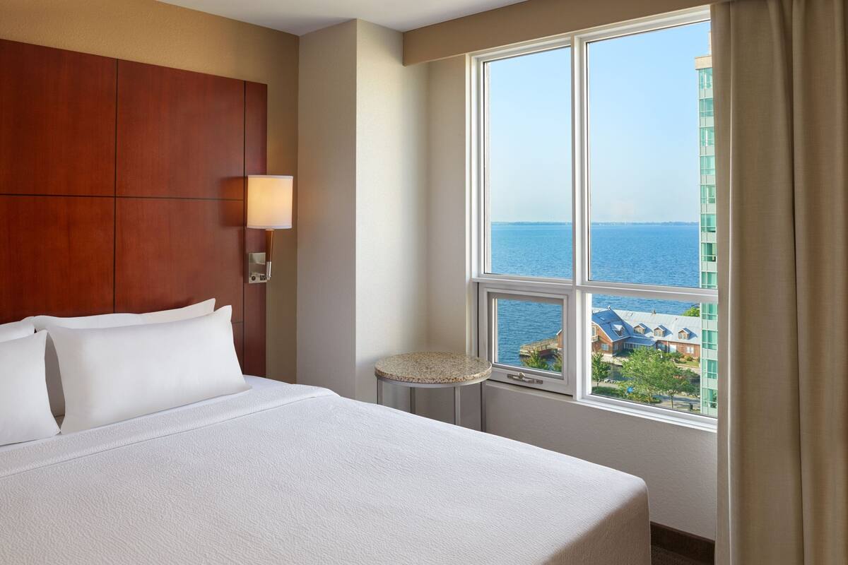 A hotel room with a large bed, a small round table, a nightstand, a lamp, and a window with a view of the sea and buildings.