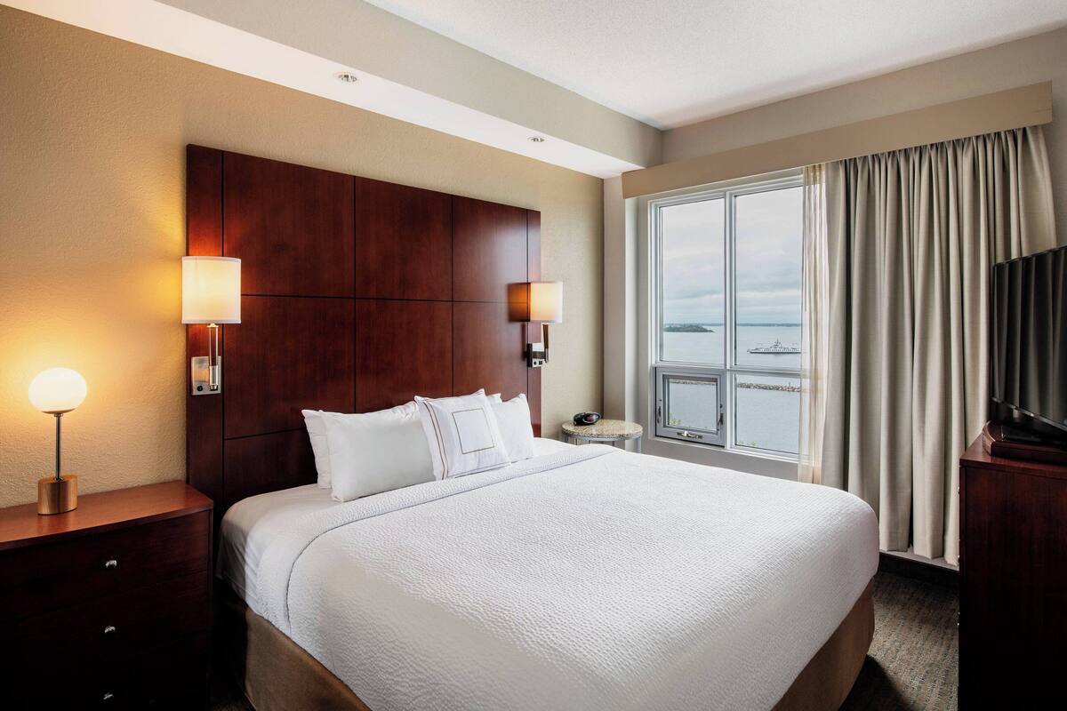 A hotel room with a large bed, two bedside lamps, a TV, and a view of the sea through a window with curtains.