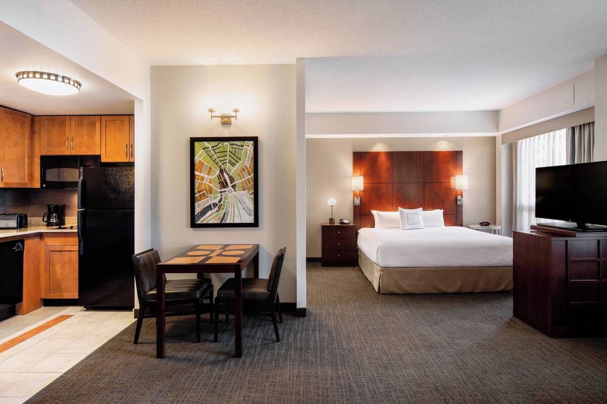 A hotel room with a bed, a TV, a desk with chairs, a kitchen area, and a wall-mounted artwork.